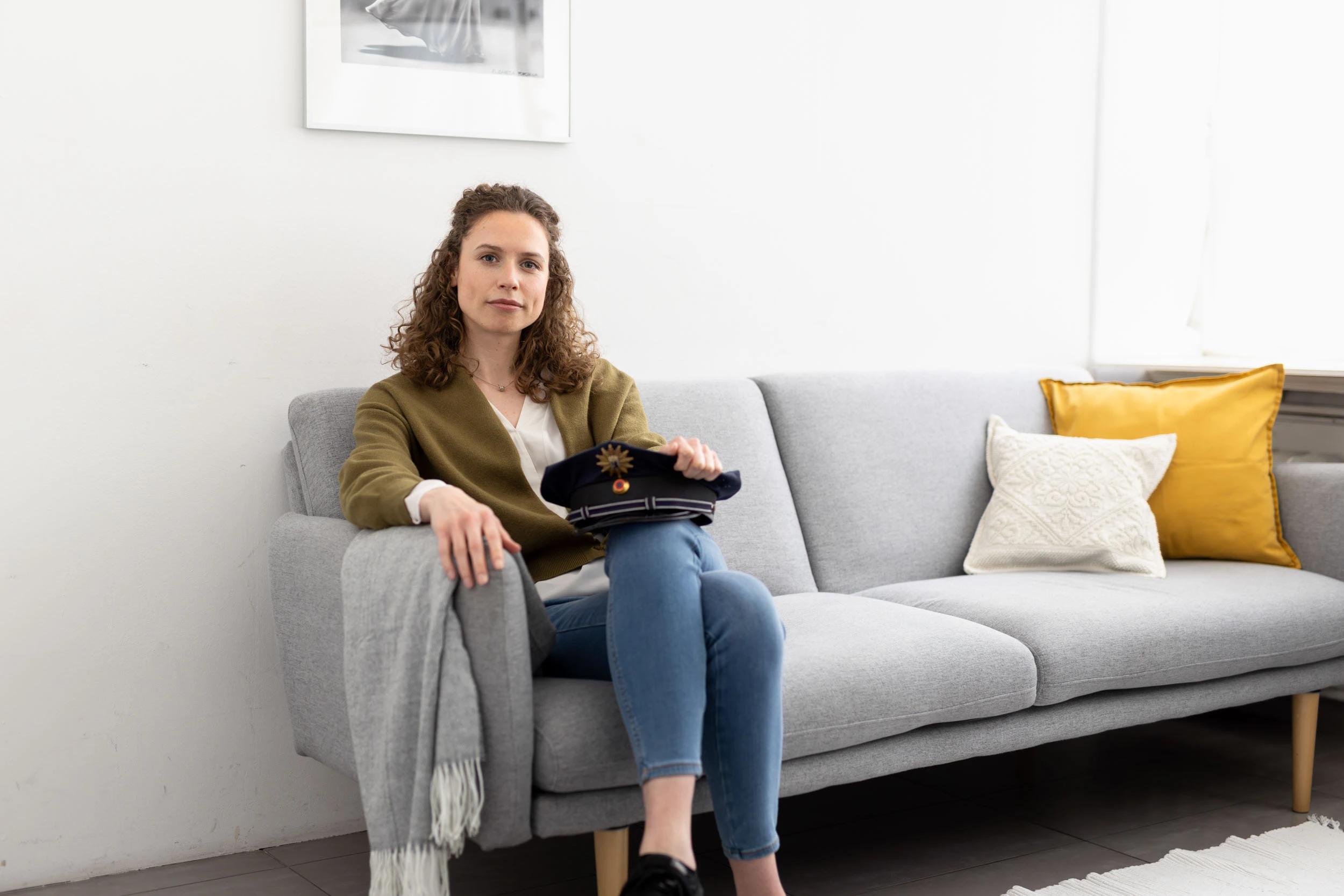Eine bei der Berliner Polizei beschäftige junge Frau sitzt lächelnd auf einem grauen Sofa in ihrem berlinovo-Apartment für Landesbeschäftigte. Auf ihrem Schoß hält sie ihre Dienstmütze.