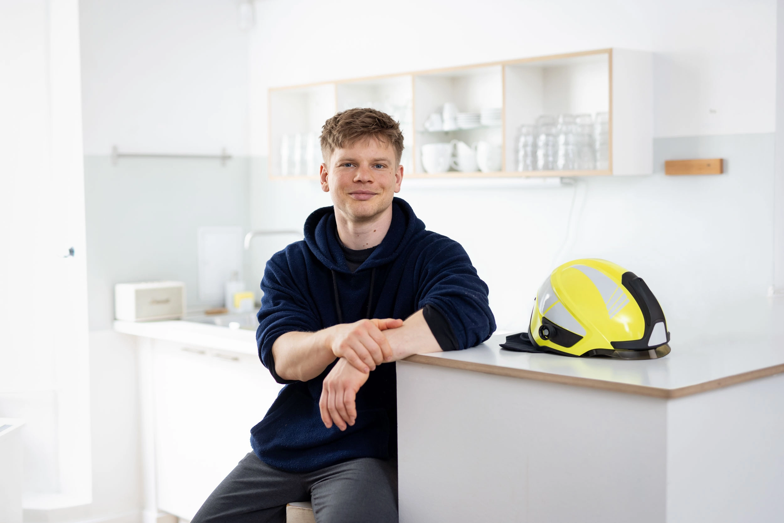 Der bei der Berliner Feuerwehr beschäftigte junge Mann sitzt in der Küche eines Apartments. Auf dem Tisch vor ihm liegt sein Feuerwehrhelm.