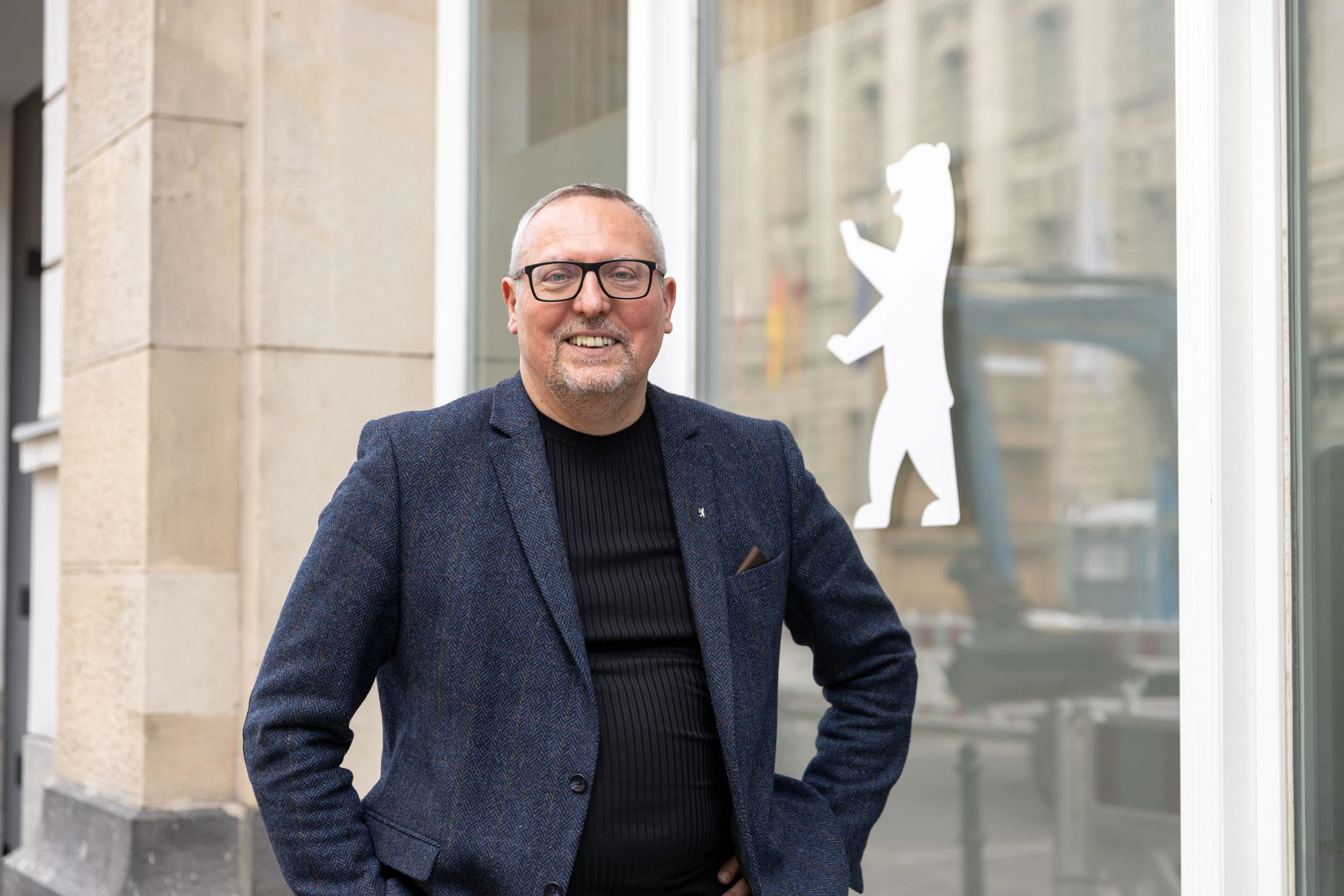 Portraitfoto von André Lossin, fotografiert vor der Senatsverwaltung für Inneres und Sport. An der Fensterfront hinter ihm ist ein silberner Aufkleber des Berliner Bären angebracht.